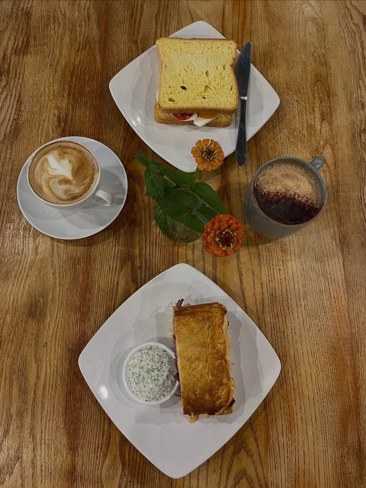 Overhead view of a Wadhams breakfast spread with two plates of toast and pastries, a latte, a mocha, and orange flowers on a wooden table