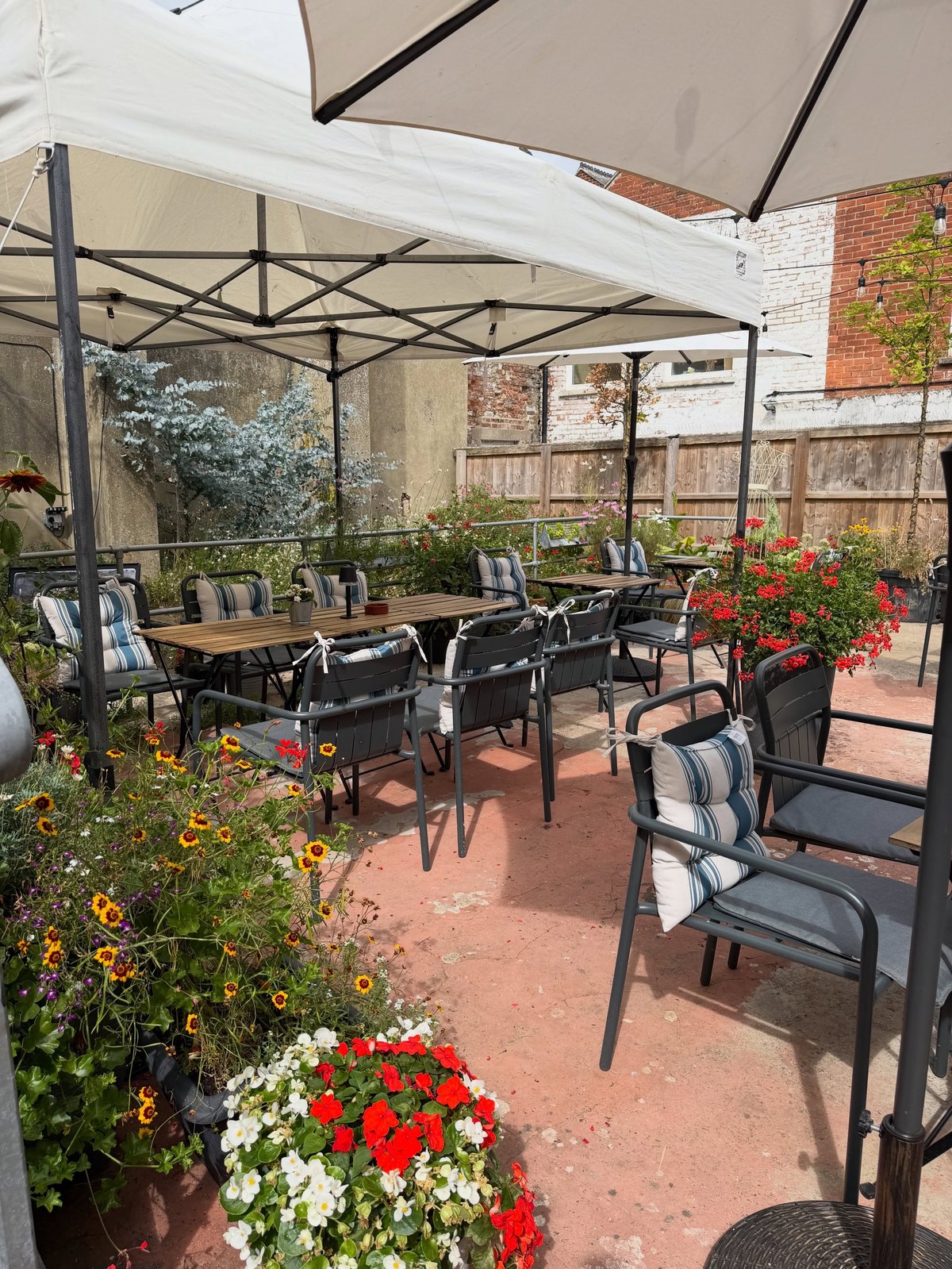 Wadhams outdoor patio with black metal chairs, wooden tables, gazebo canopy, brick walls and red and white flowering plants